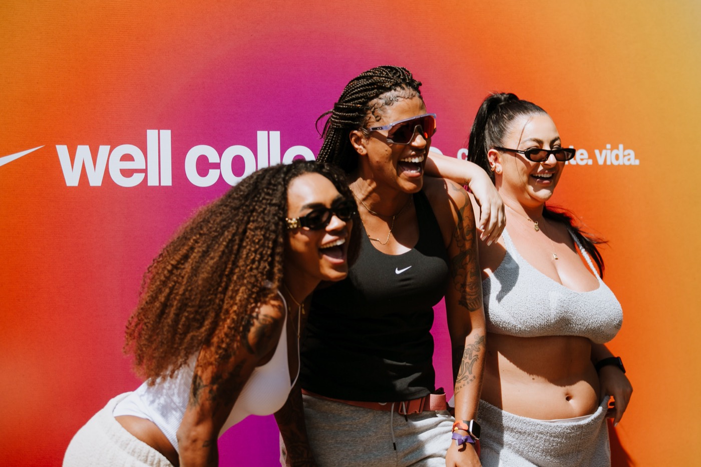 Three people in sunglasses laugh together in front of an orange and pink gradient panel on a sunny day