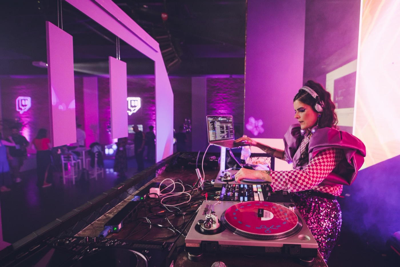 DJ in headphones and a pink checkered outfit plays on turntables, with the hall in purple light behind