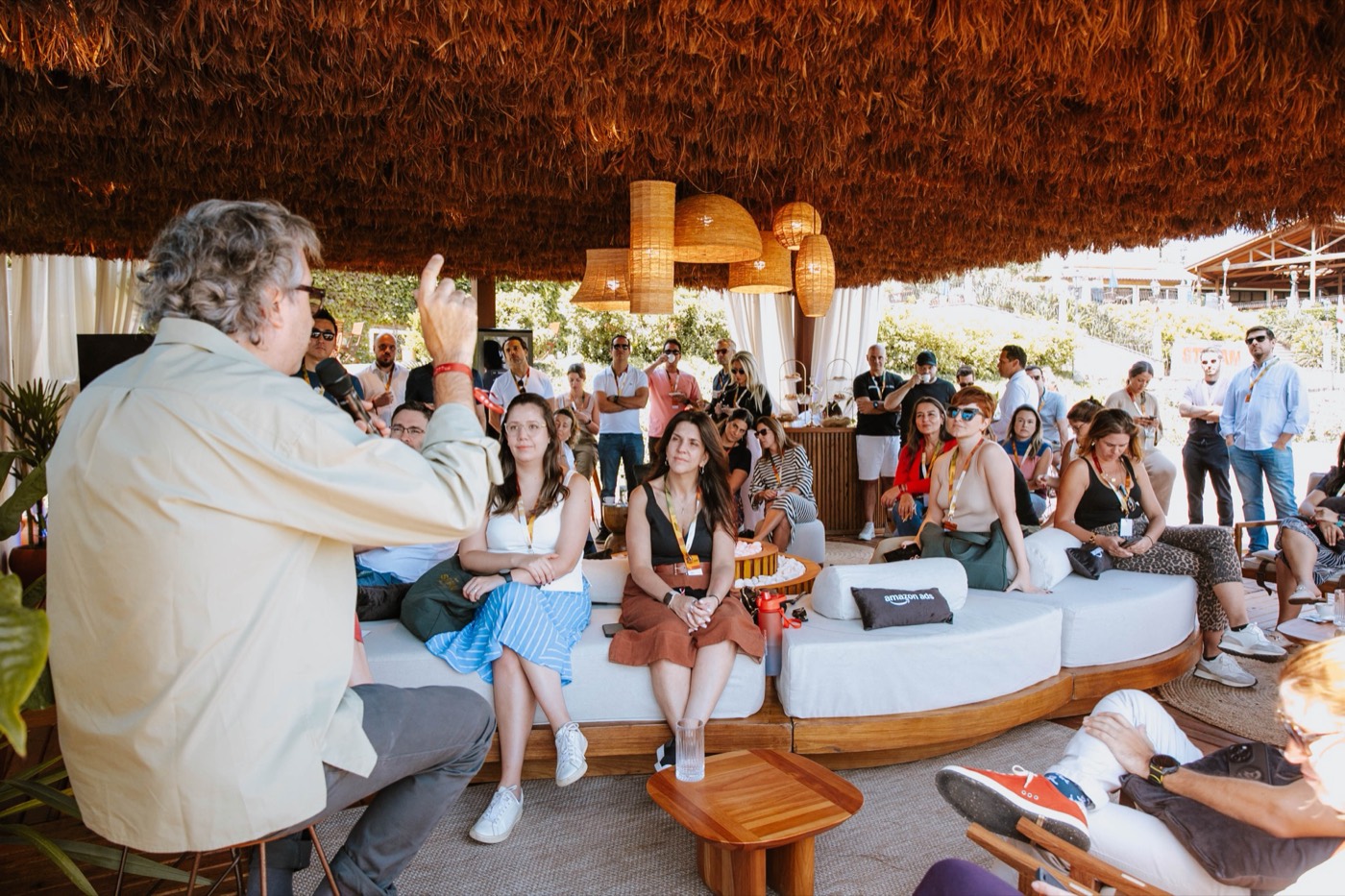 Person seen from behind speaks to a group seated on white sofas under a thatched roof with pendant lamps