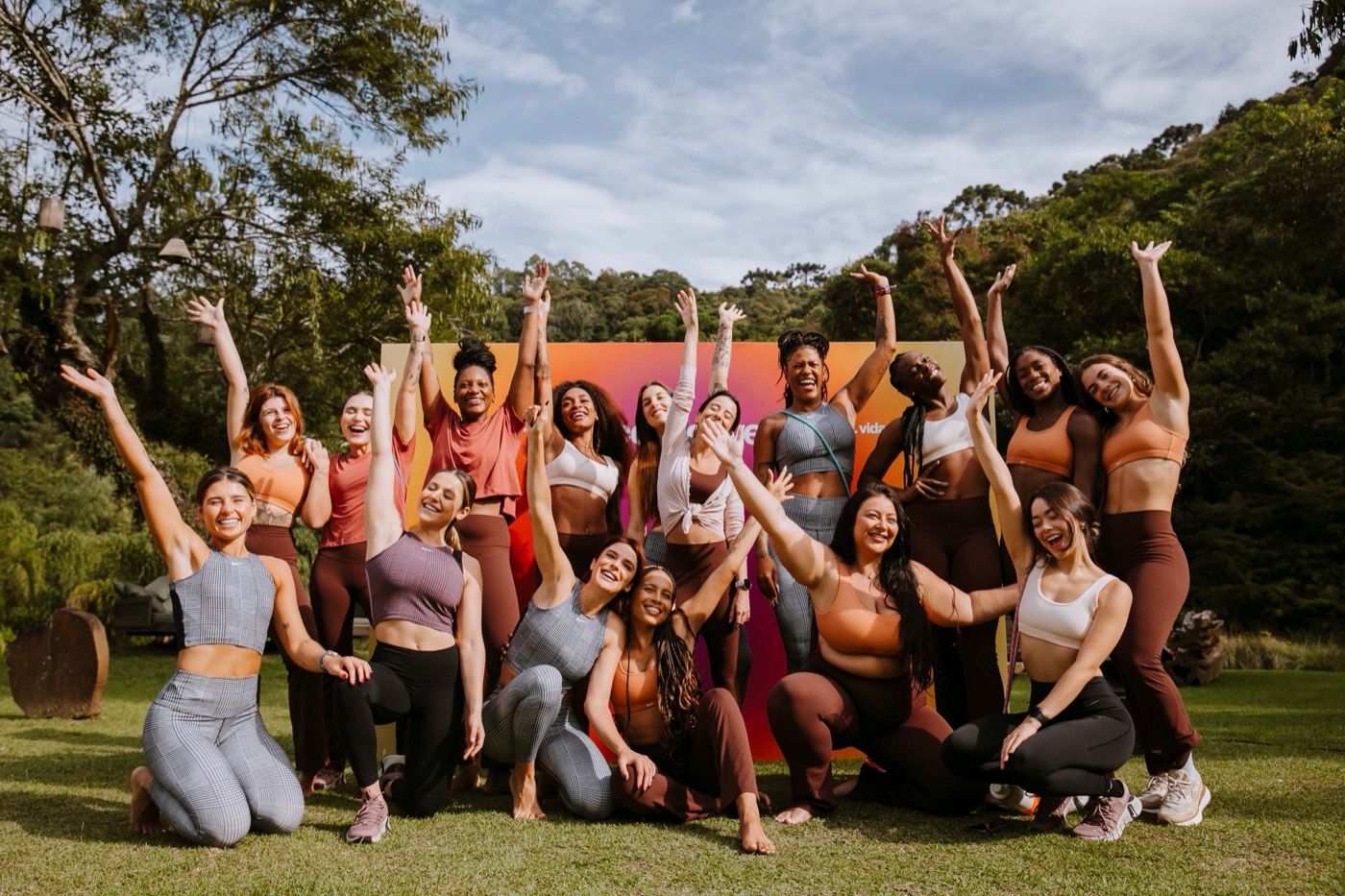 Large group poses with arms raised on a lawn, in front of a colorful panel and surrounded by trees