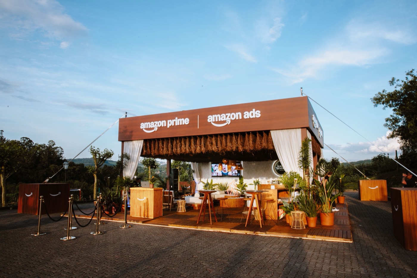 Outdoor wooden booth with "amazon prime" and "amazon ads" on the fascia, white curtains, plants and rope stanchions