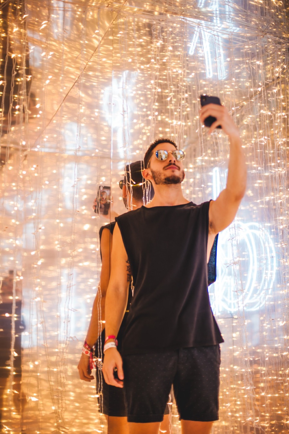 Person in mirrored sunglasses takes a selfie with a phone in a mirrored room with golden string lights