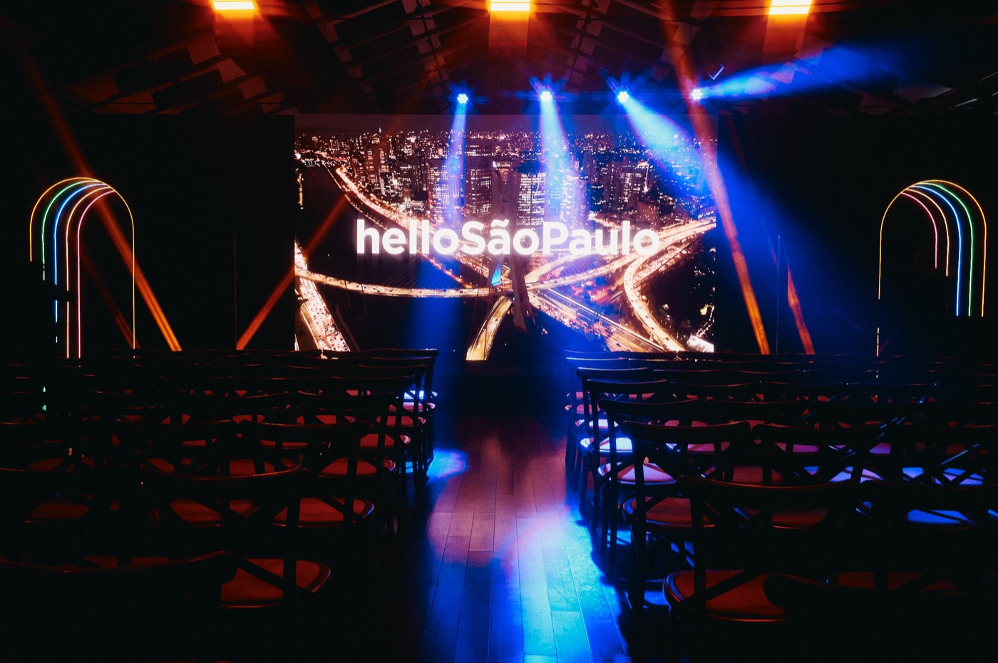 Empty seating and center aisle facing a screen reading "helloSãoPaulo" under blue light beams