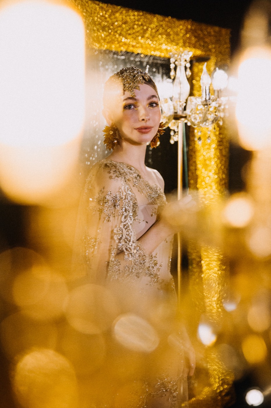 Person in a gold-embroidered dress in front of a glittering gold frame, seen through blurred lights