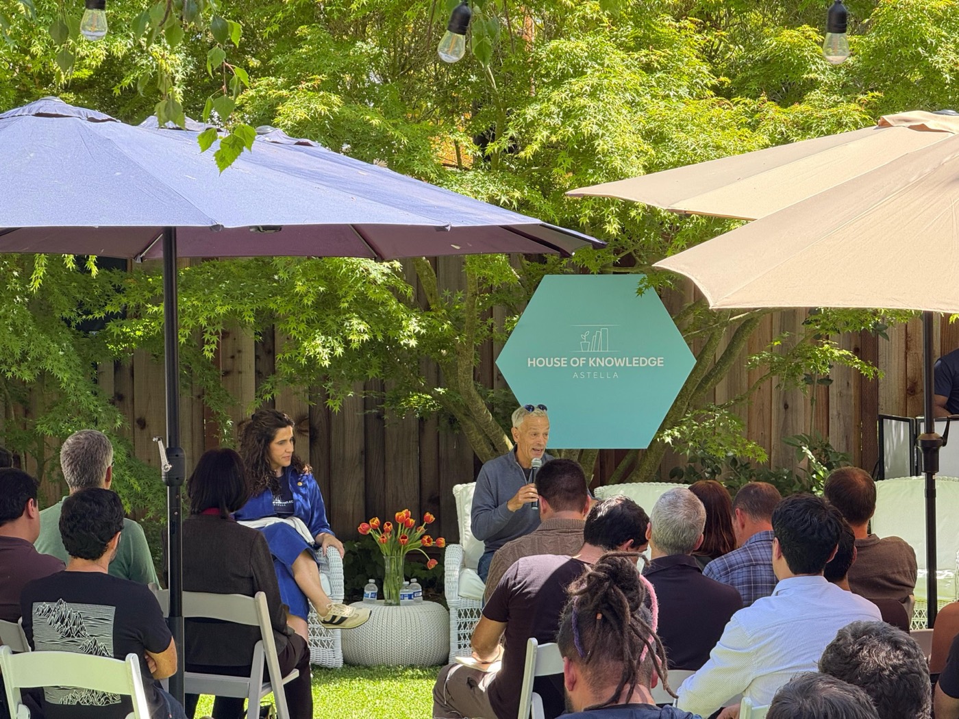 Two people seated facing the audience in a garden, one of them speaking into a microphone, under umbrellas