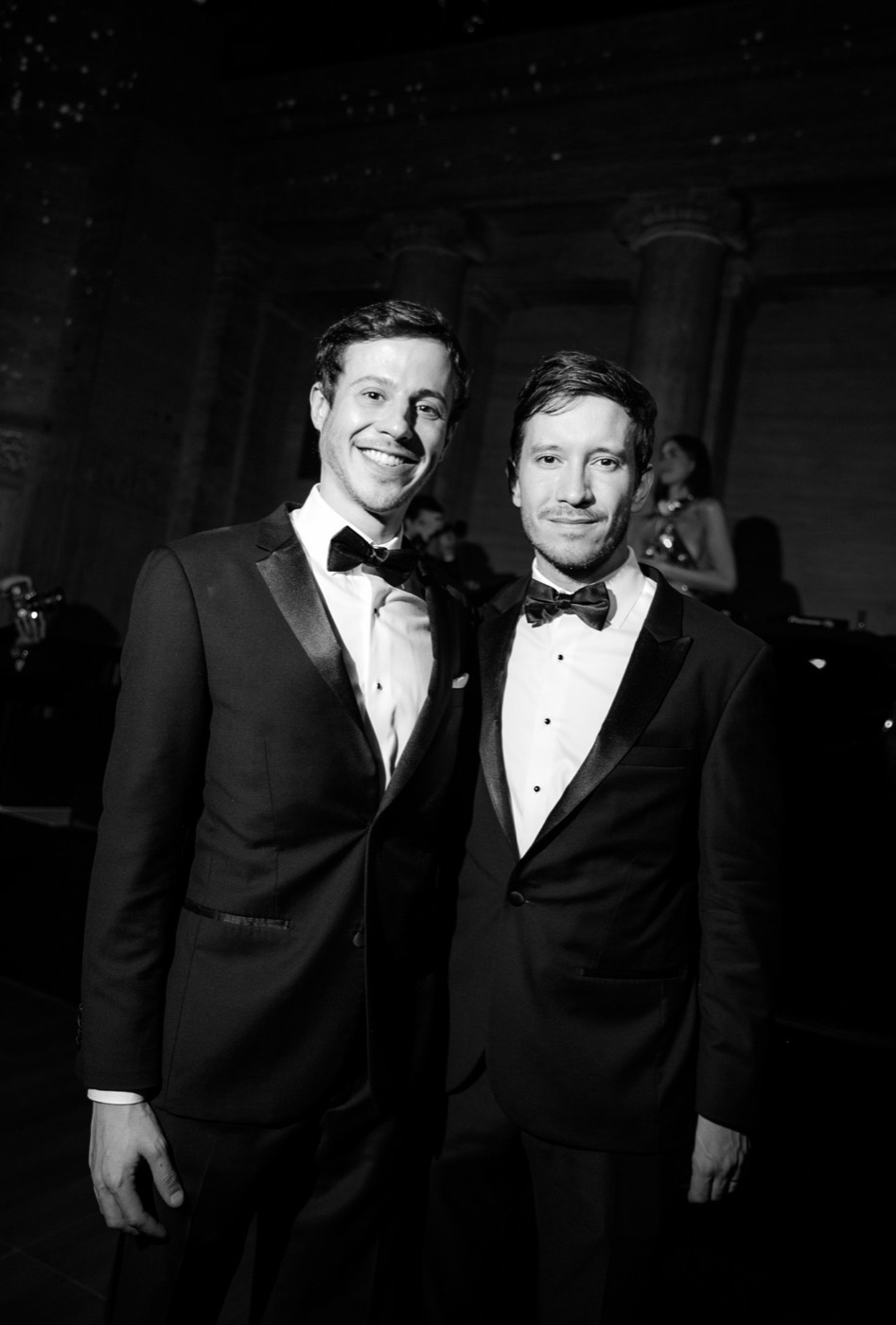 Two people in tuxedos and bow ties pose smiling side by side in a dark hall, in black and white