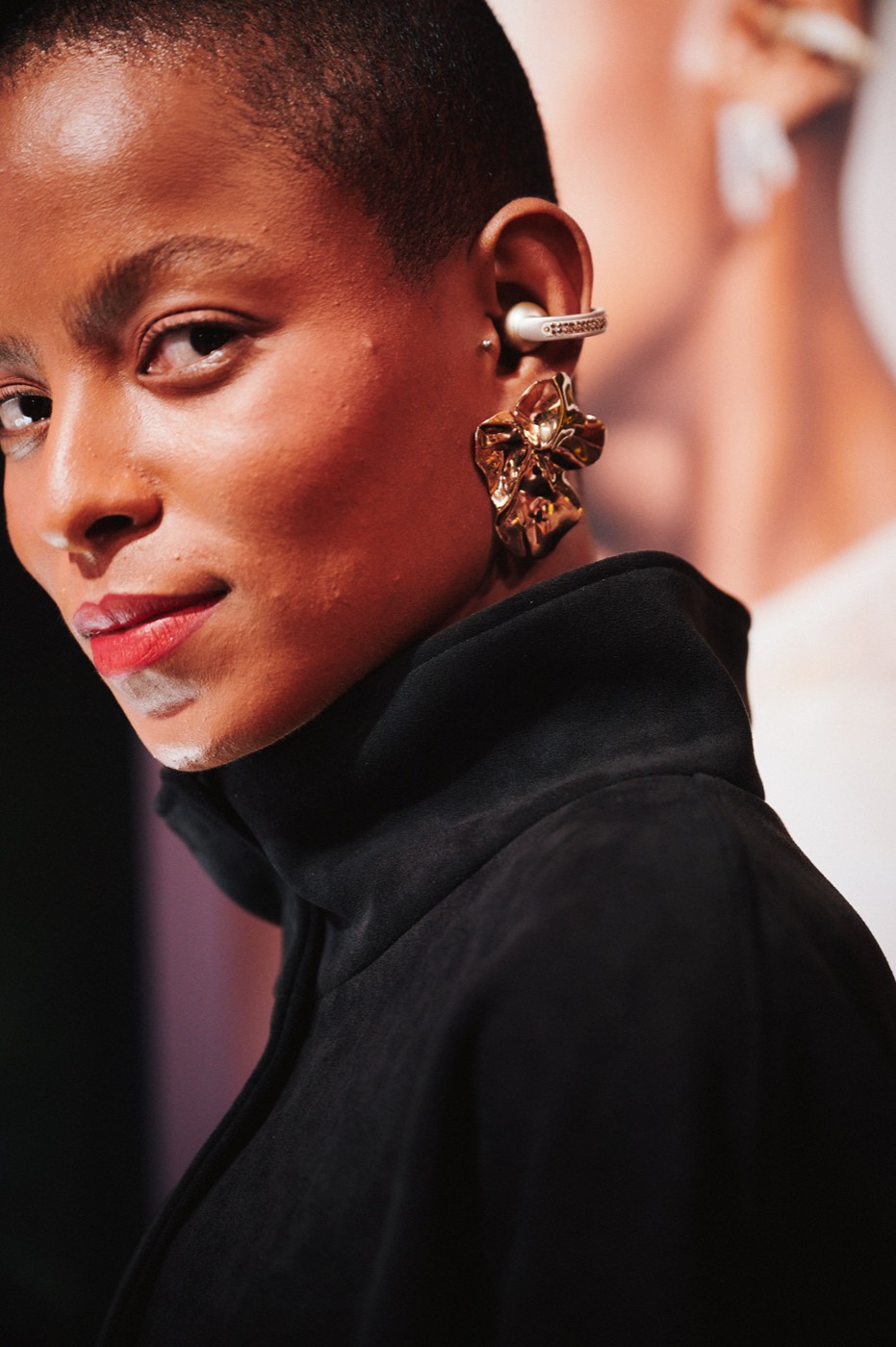 Close-up of a person with a shaved head, a gold flower-shaped earring and black clothing