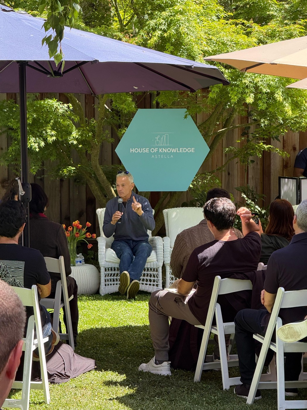 Person seated in a white armchair speaks into a microphone to an audience on folding chairs on the lawn