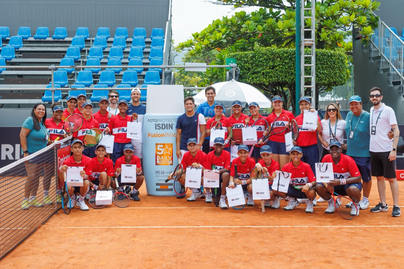 Large group in red shirts poses on a clay court around a giant "ISDIN" bottle replica