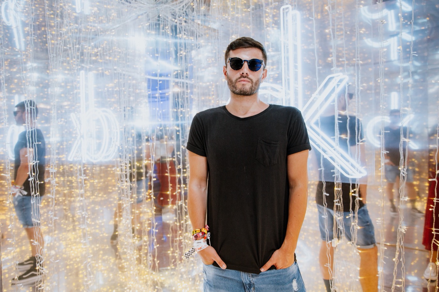 Person in sunglasses and a black T-shirt poses before a neon sign in a mirrored room with string lights