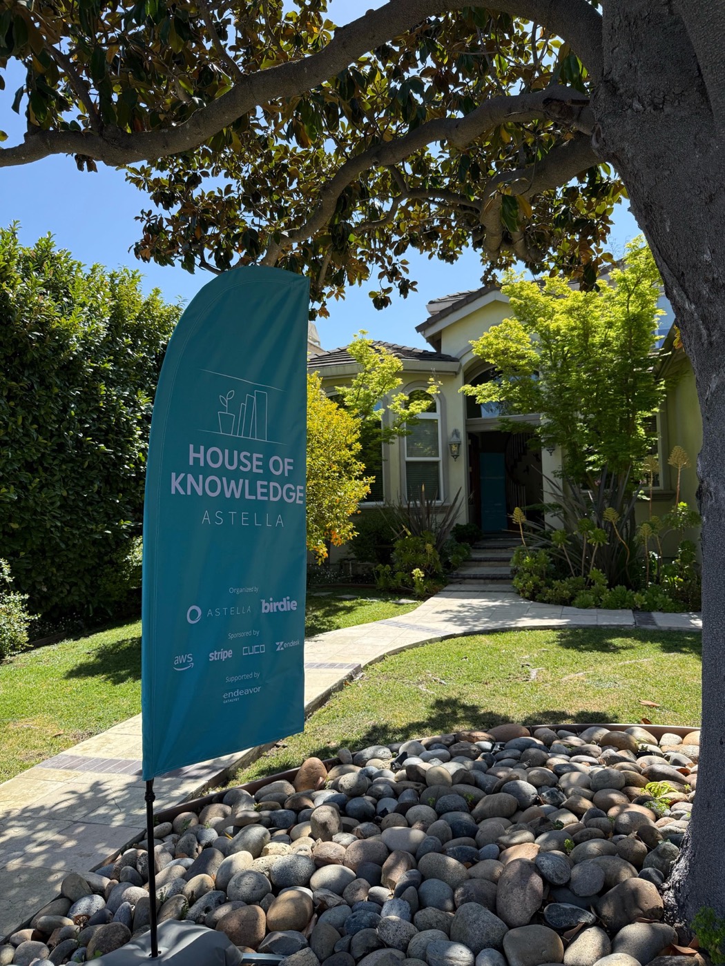 Teal vertical flag banner reading "HOUSE OF KNOWLEDGE ASTELLA" in a garden with river stones, in front of a house