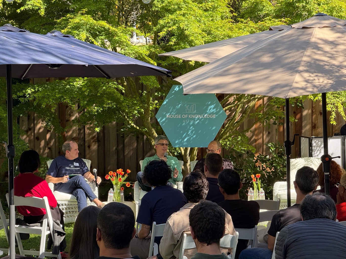 Wider view of the garden talk, with three people seated under umbrellas and the audience seen from behind