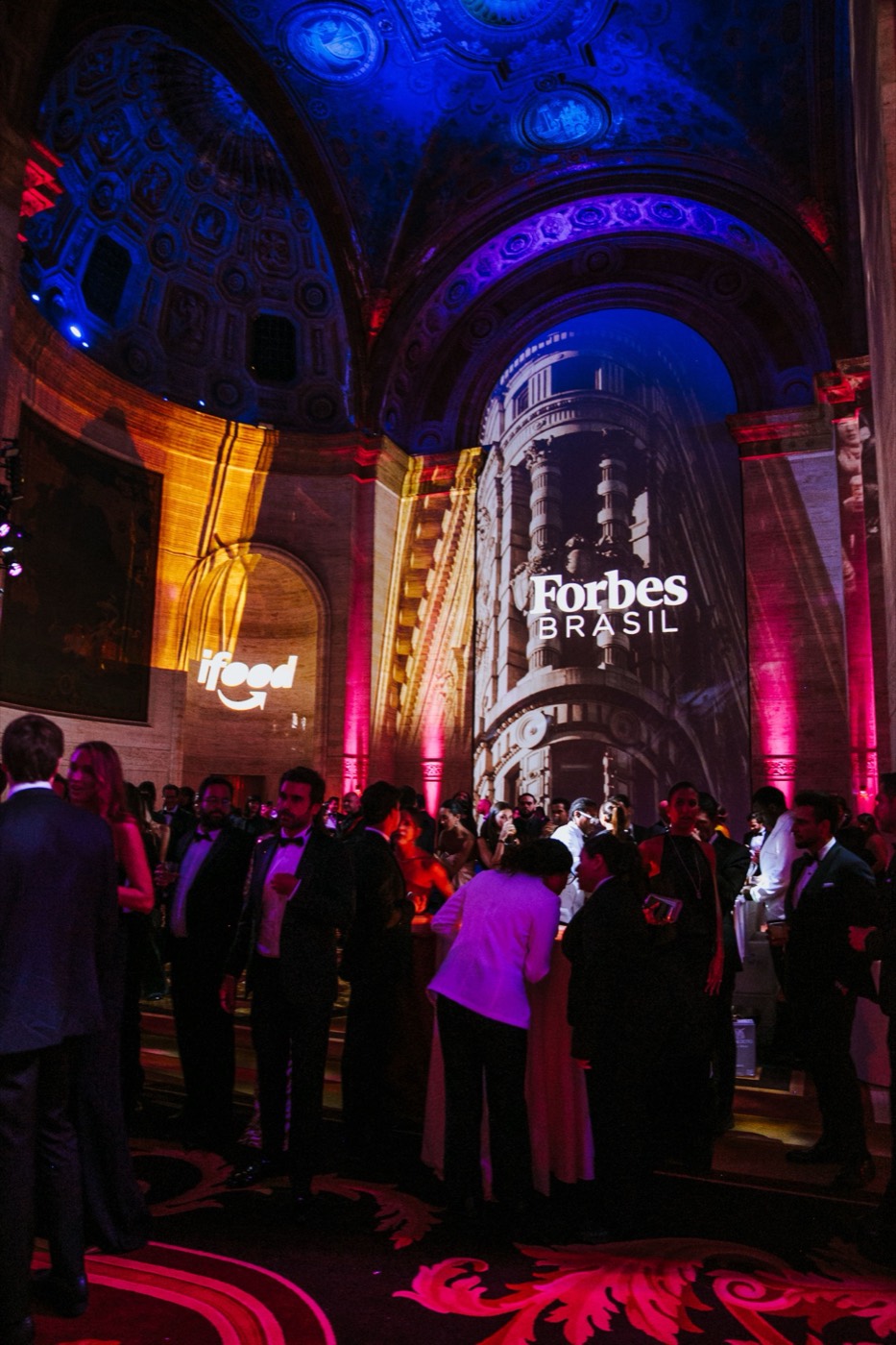 Guests in formal wear beneath a vaulted ceiling with a "Forbes BRASIL" projection and pink and blue lights