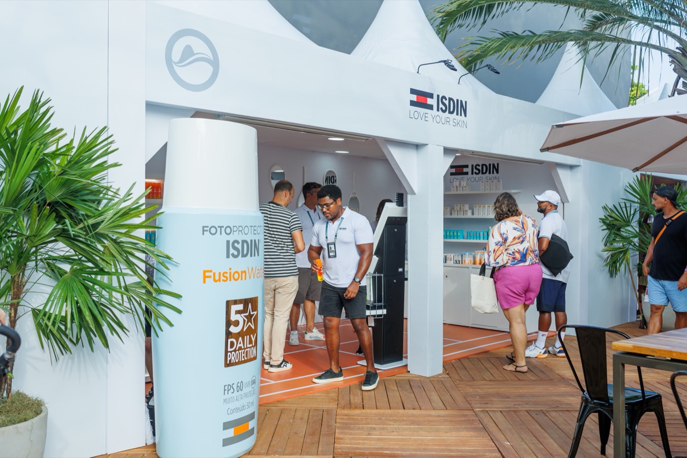 White "ISDIN" booth with a giant bottle replica at the entrance, a wooden deck and visitors walking around