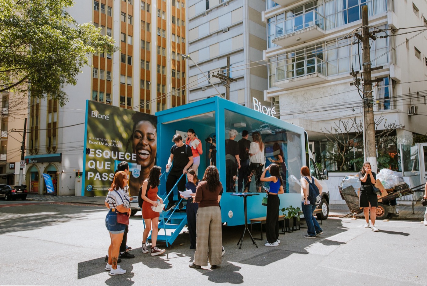 People line up and move around an open blue "Bioré" truck on a street with buildings