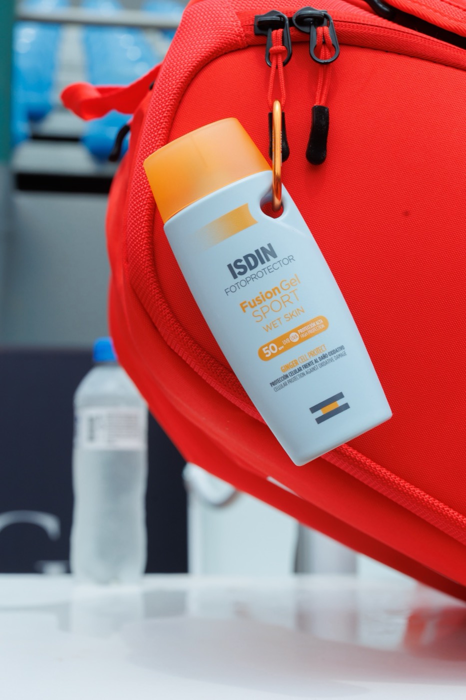 "ISDIN" sunscreen bottle clipped with a carabiner to a red sports bag, with a water bottle in the background