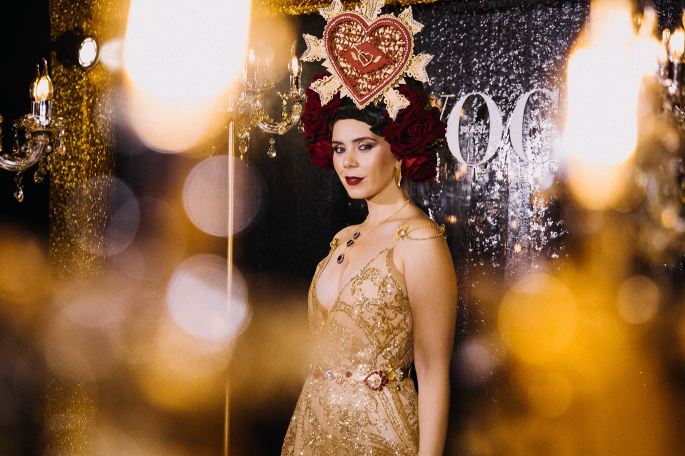 Person wearing a gold heart and red flower headpiece and a gold dress, among blurred lights
