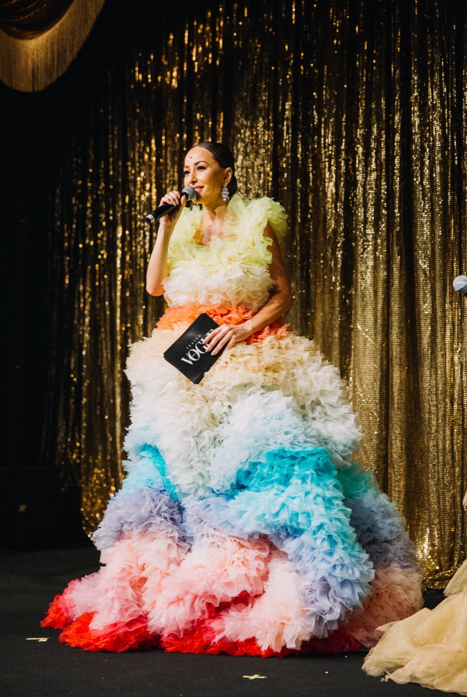 Person on stage with a microphone in a voluminous tulle gown in colorful layers, in front of a gold curtain