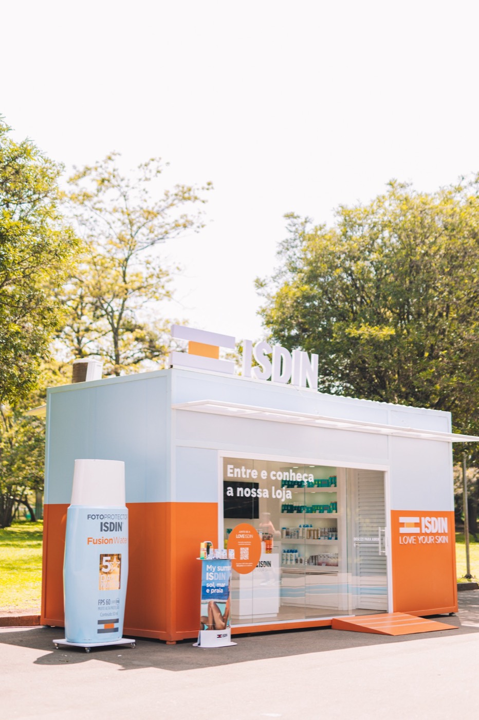 White and orange container store with "ISDIN" signage in a tree-lined area, with a giant bottle replica beside it