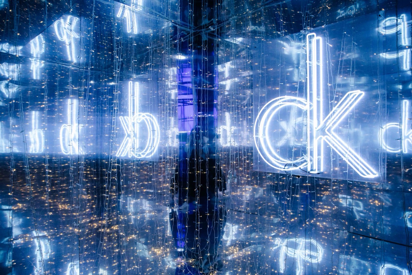 Mirrored room in blue light with a neon "ck" sign reflected many times and strands of string lights