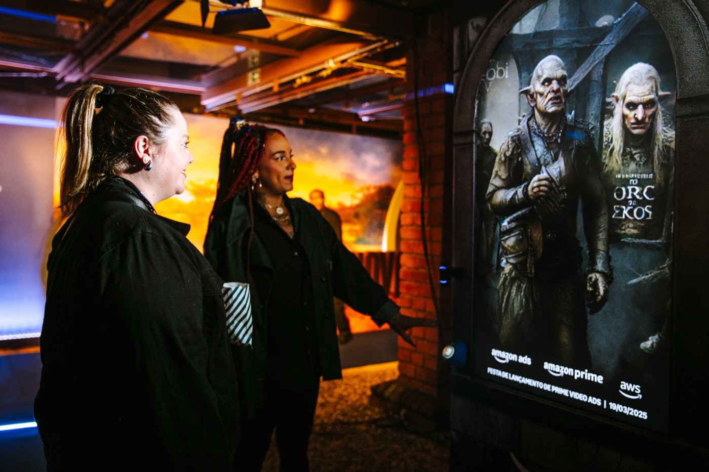 Two people look at a backlit poster with fantasy characters and "amazon ads", "amazon prime" and "aws" logos