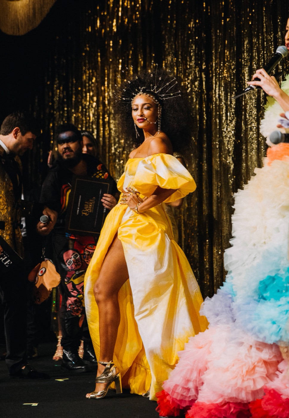 Person in a yellow slit dress and a spiked headpiece poses on stage in front of a gold curtain