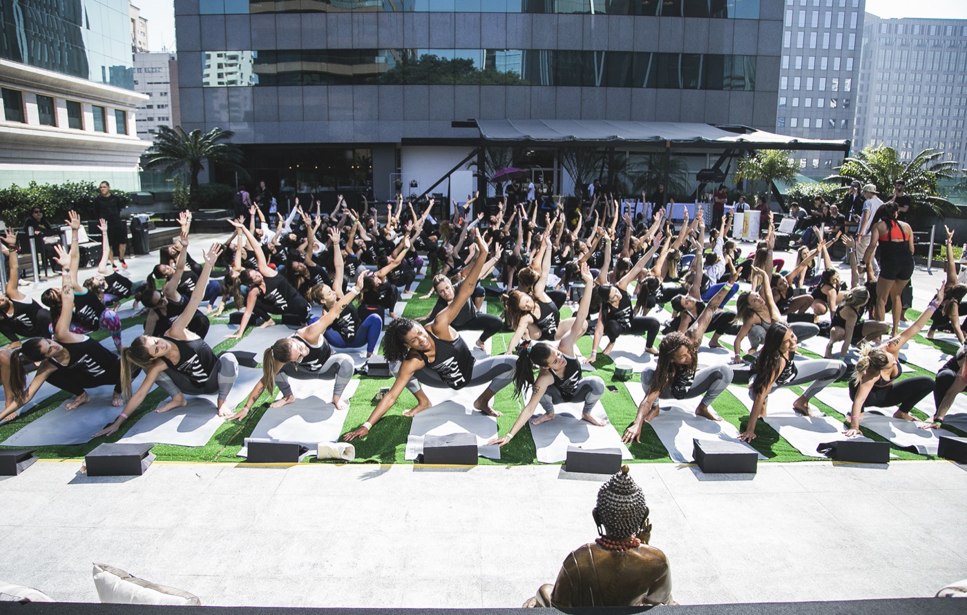 Large group on mats outdoors stretches one arm to the side, with a Buddha statue in the foreground