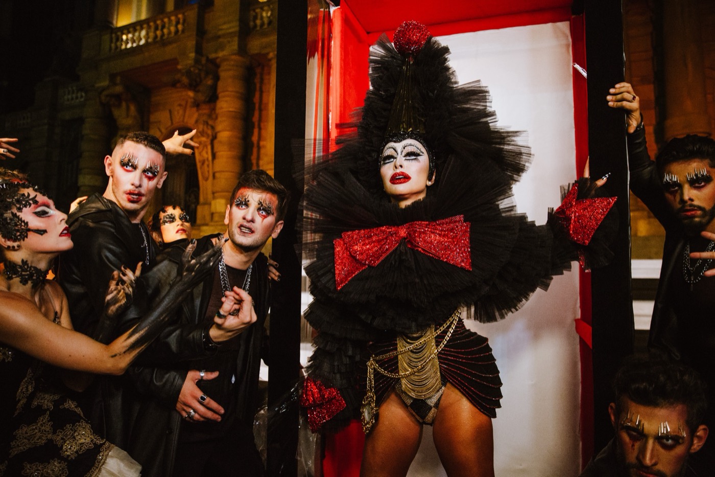 Person in a voluminous black costume and sparkling red bow in a red booth, surrounded by people in theatrical makeup