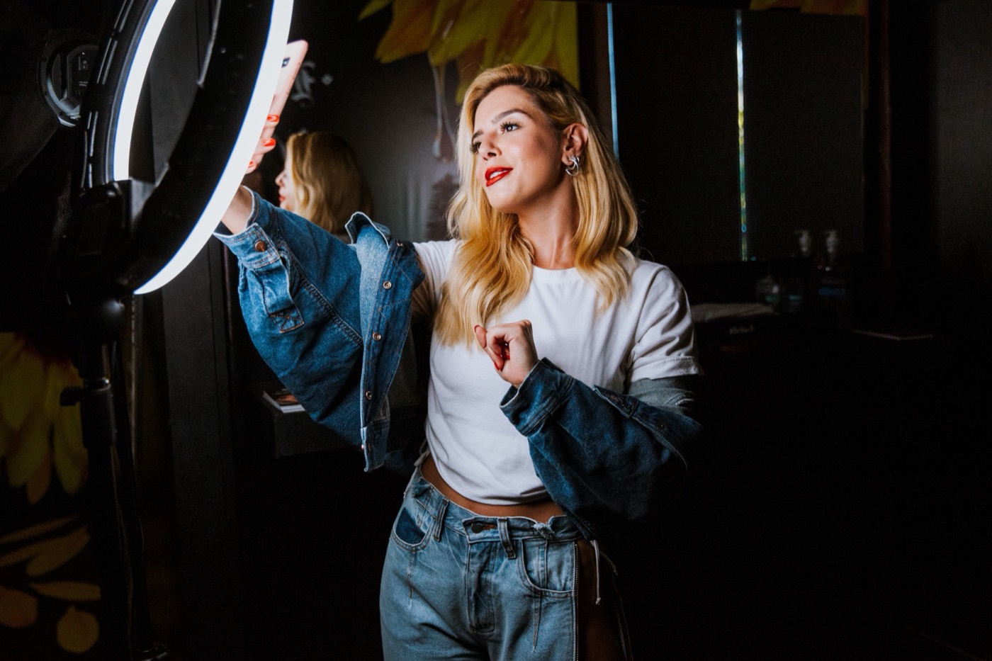 Person in a denim jacket takes a phone selfie in front of a ring light in a dark room
