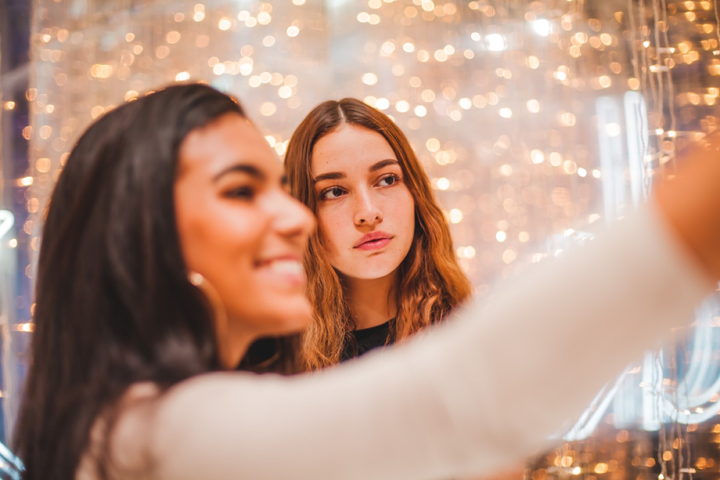 Two people take a selfie in the foreground against a blurred curtain of string lights