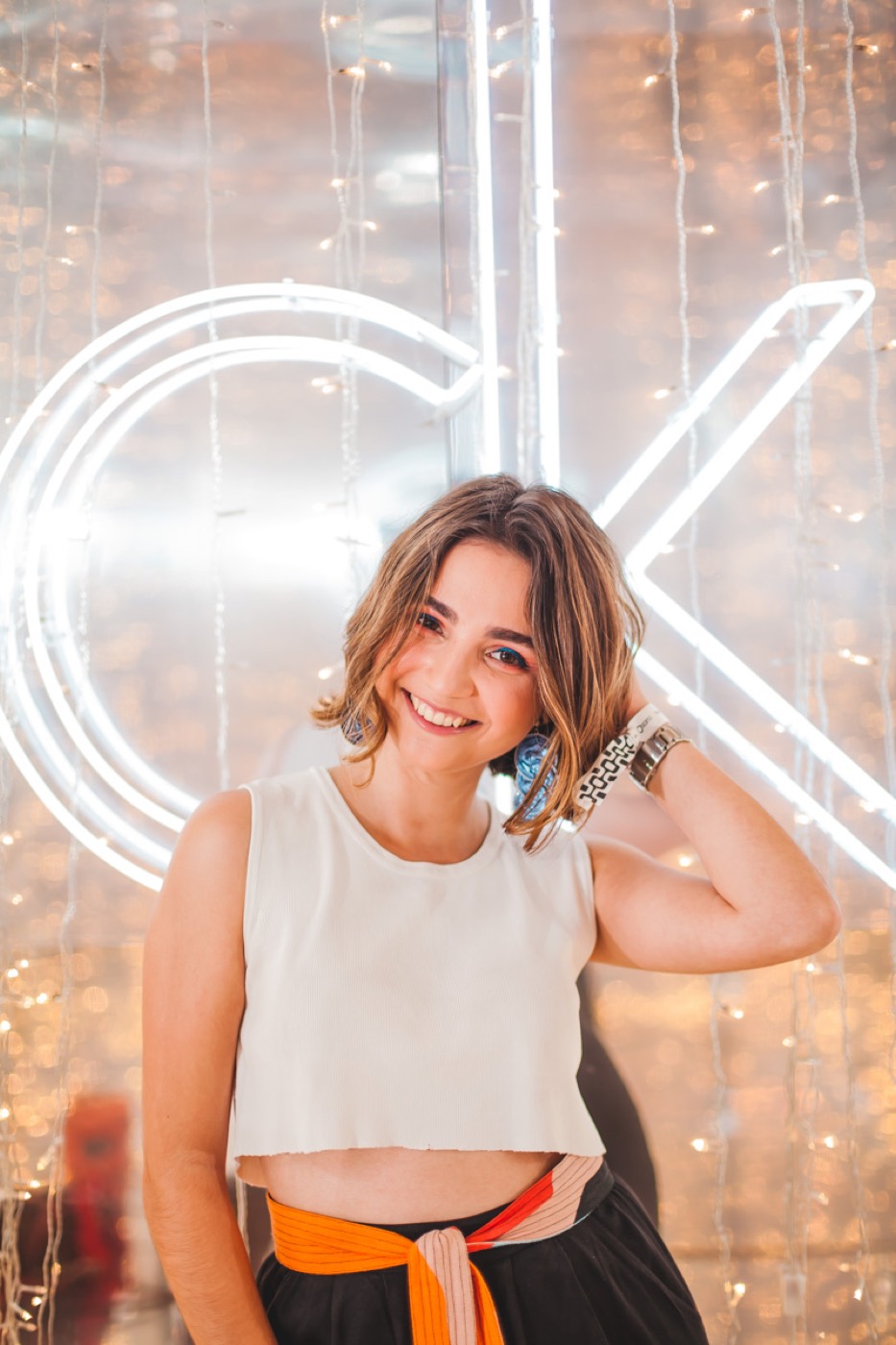 Person with short hair smiles, hand behind the neck, in front of a neon "ck" sign and string lights