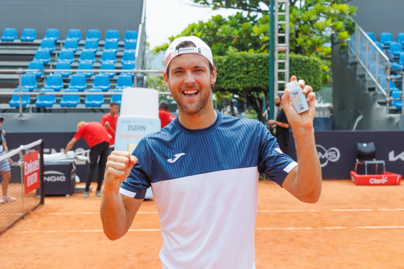 Person in a cap and sports shirt smiles on a clay court holding a bottle, with blue stands behind