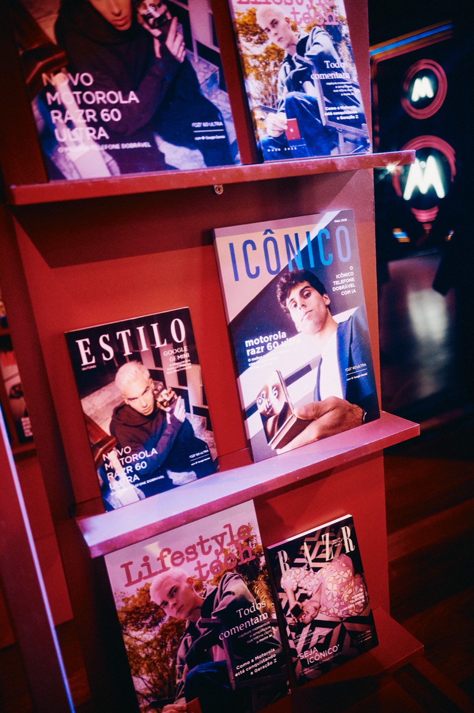 Red shelves with magazines whose covers read "ESTILO", "ICÔNICO" and "Lifestyle tech", with pink neon in the background