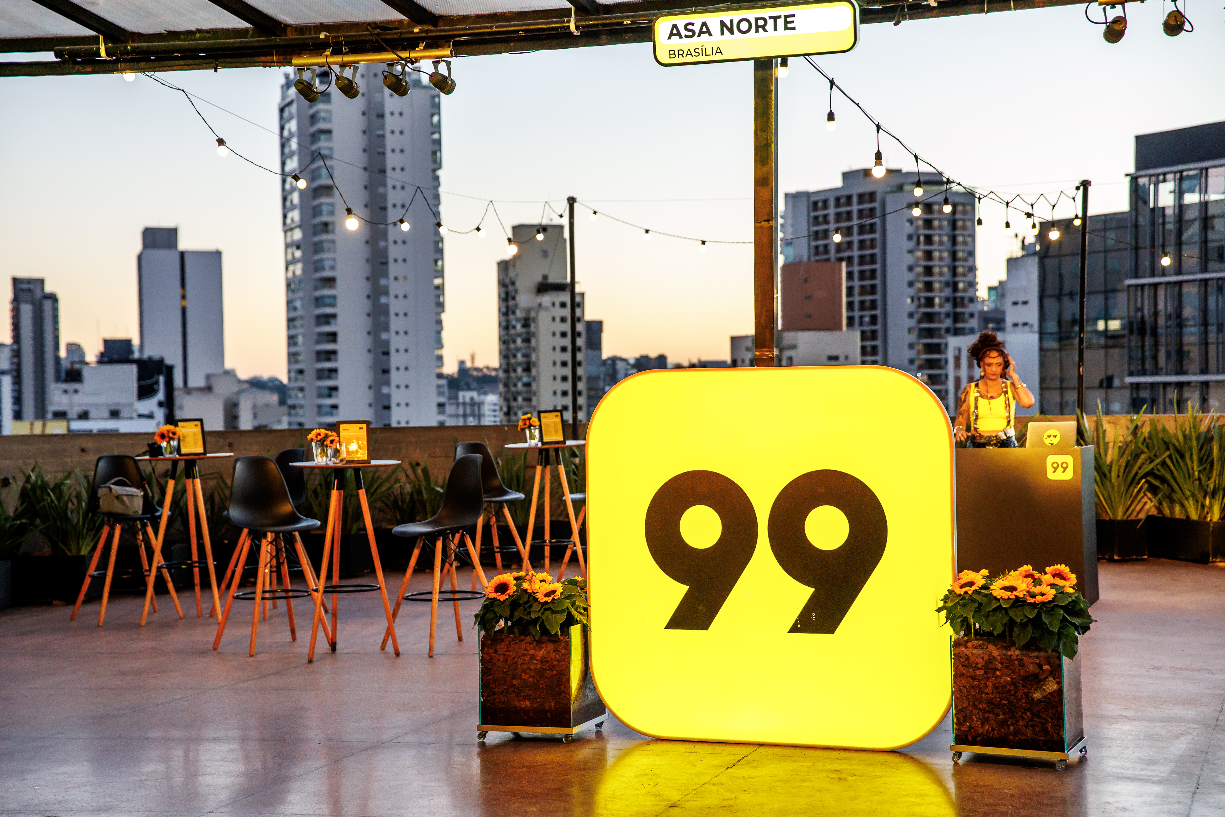Terrace at dusk with a glowing "99" totem, high tables, flower pots and a person at a DJ booth