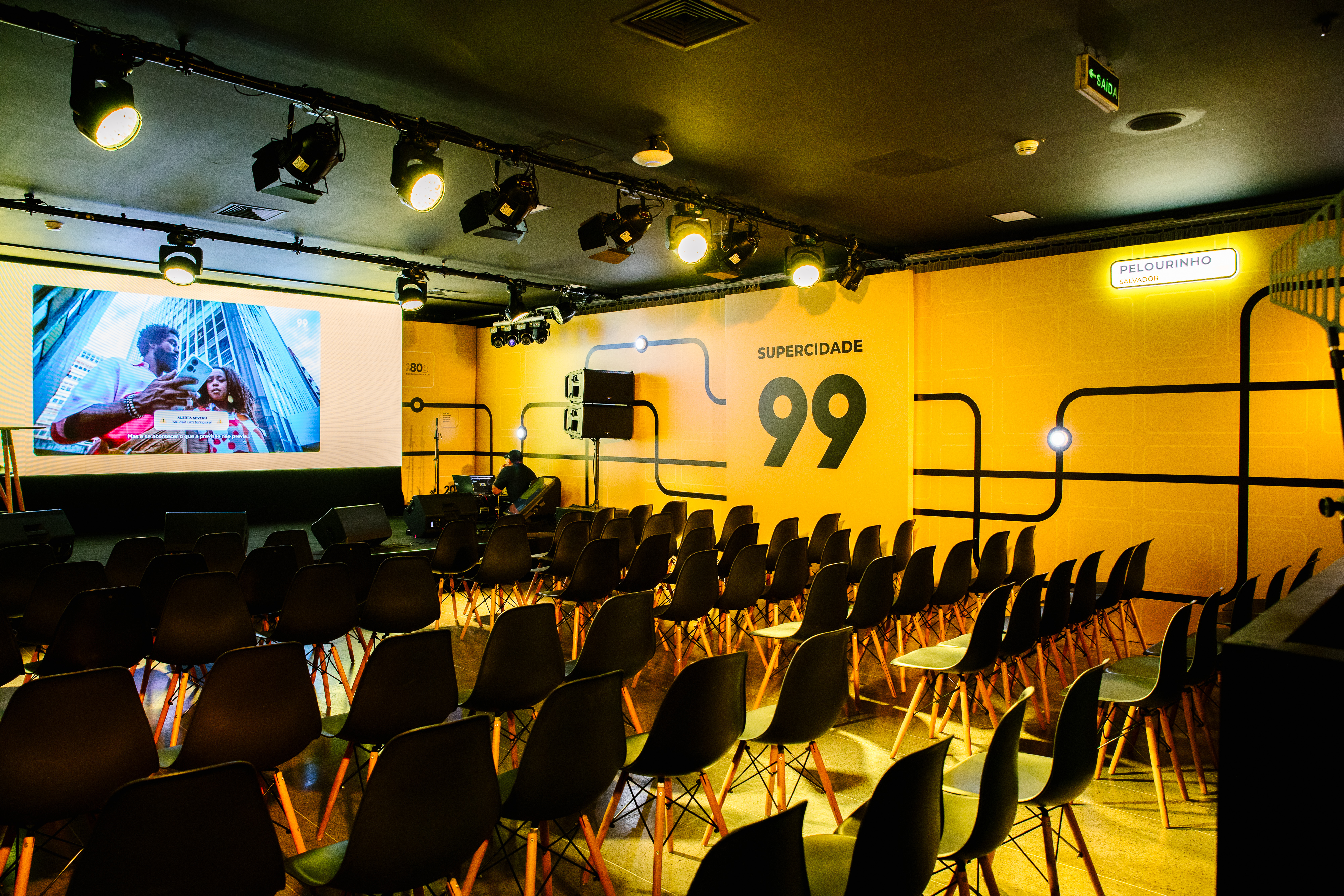 Auditorium with rows of black chairs, a screen and a yellow wall with "SUPERCIDADE 99" lettering