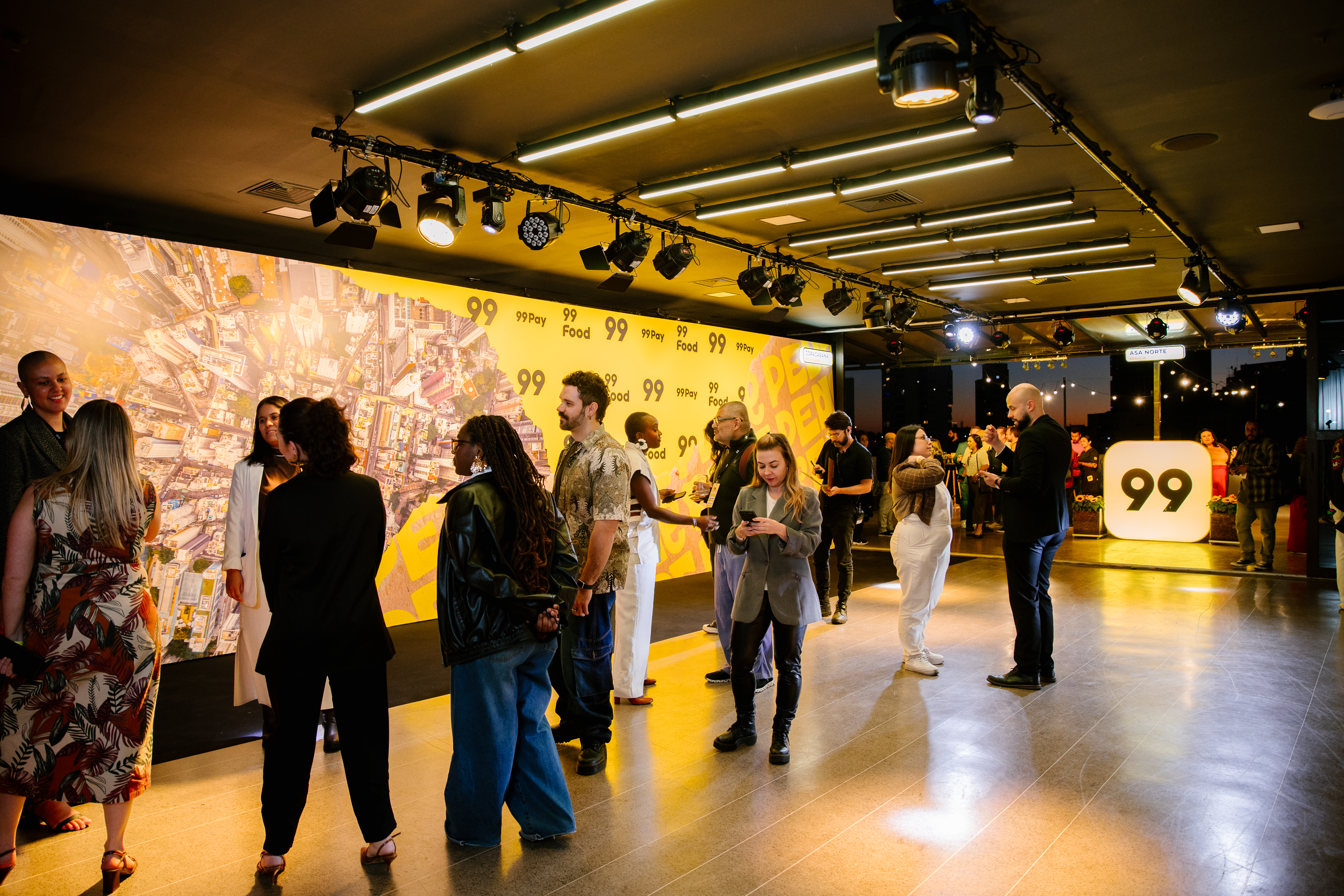 Guests chat in a hall in front of a panel with an aerial map and "99" logos