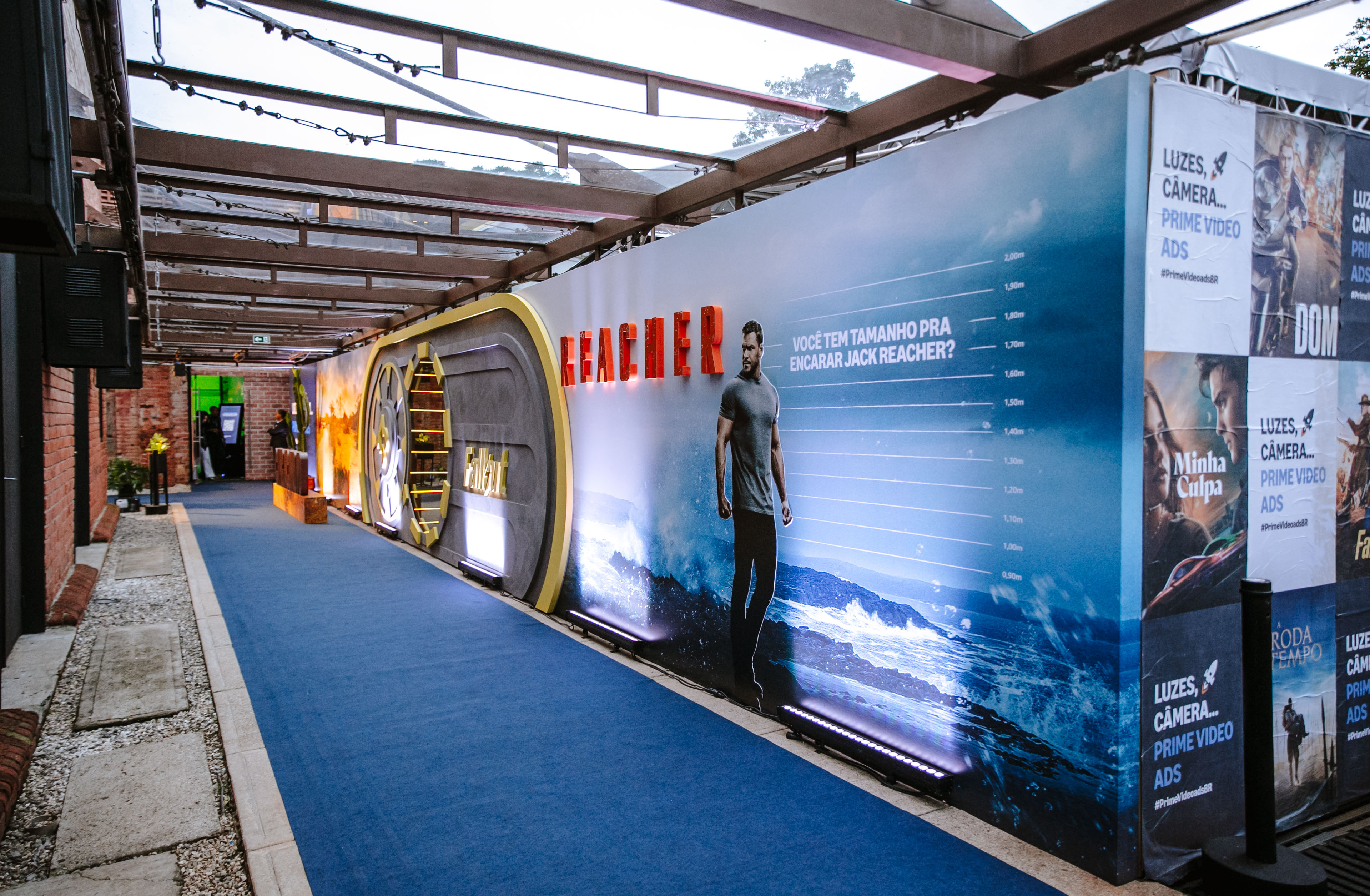 Outdoor walkway with blue carpet and series panels, with "REACHER" lettering and "PRIME VIDEO ADS" posters