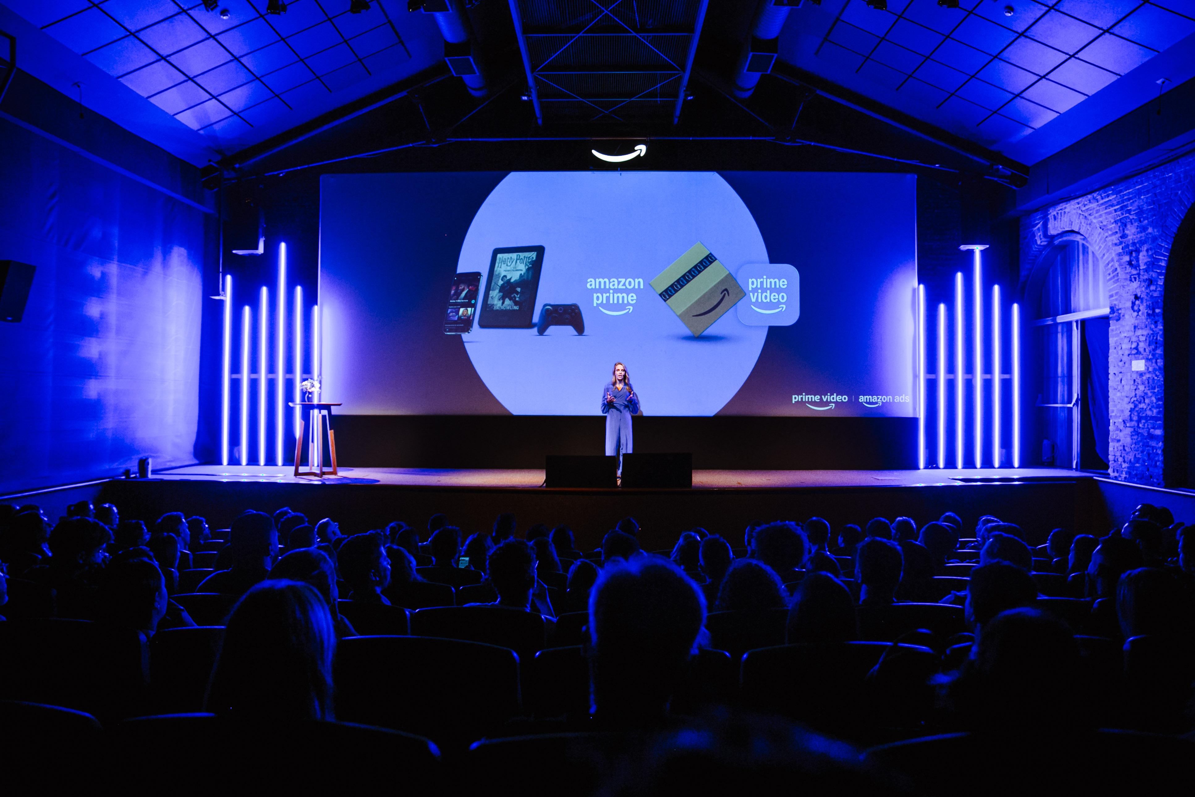 Person on a blue-lit stage in front of a screen with "amazon prime" and "prime video" logos, audience in silhouette