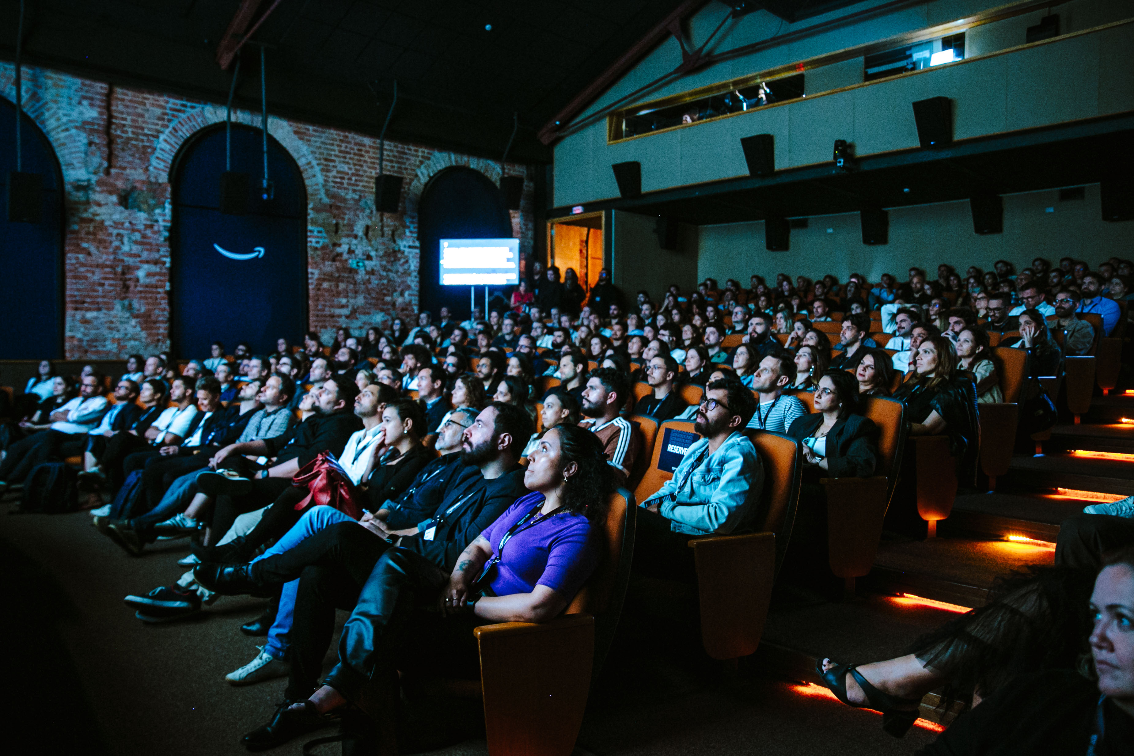 Audience seated in an auditorium with upholstered seats and brick walls watches the presentation